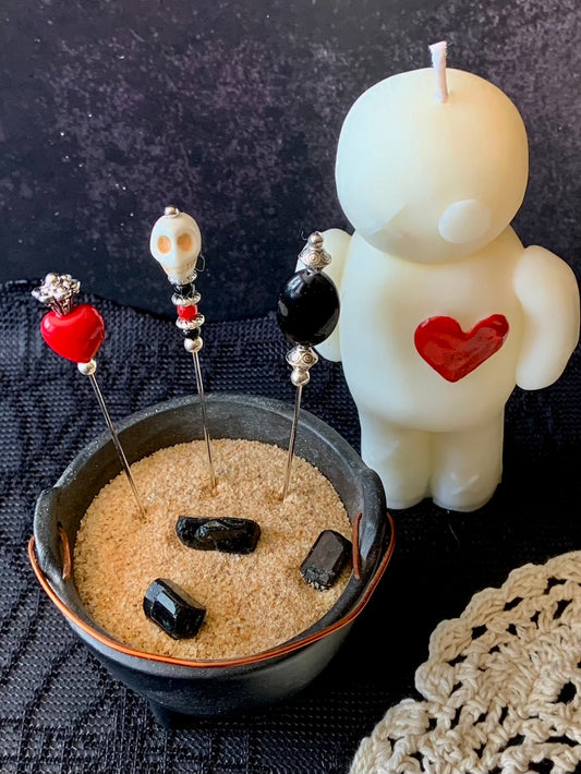 Decorative items including a white candle with a red heart, black stones, and decorative sticks in a bowl on a dark surface.